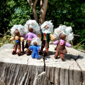 May include: Five handmade felt dolls with white hair and painted faces. The dolls are wearing colorful clothing and are standing on a wooden surface.