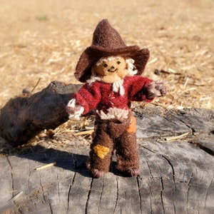 May include: A handmade felt scarecrow doll with a brown hat and pants, a red shirt, and straw hair. The doll has a stitched face and patches on the pants. It is standing on a weathered wooden surface.