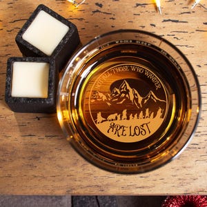 May include: A whiskey glass with the etched quote "NOT ALL THOSE WHO WANDER ARE LOST" and a mountain scene. Two square, dark gray soapstone whiskey stones with white centers sit nearby. The glass is filled with amber liquid.