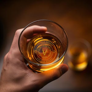 May include: A close-up of a hand holding a clear glass with amber liquid. The glass has a gold sticker on the bottom that says "Vintage Brand" with a stylized "V" in the center.