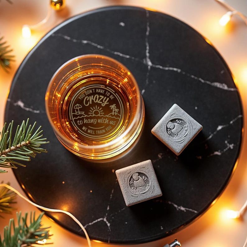 May include: A glass with a humorous saying etched on it, "You don't have to be crazy to hang with us, we will train you". The glass is filled with a brown liquid and is sitting on a black marble surface with two gray square coasters.