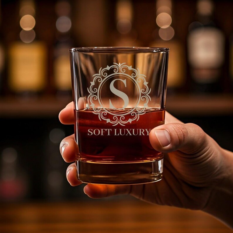 May include: A clear glass rocks glass with an etched design of a monogram "S" inside a decorative frame. The text "SOFT LUXURY" is etched below the monogram. The glass is filled with a dark amber liquid.