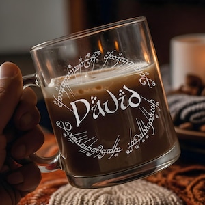 May include: A clear glass mug with a brown liquid inside. The mug has white lettering that reads "Däwid" and a circular design of Elvish script around the outside.