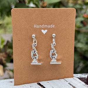 May include: Silver-colored microscope earrings displayed on a brown card. The card has the word "Handmade" and a heart symbol. The earrings are dangling and detailed, with a post at the top. The background is blurred, with green foliage.