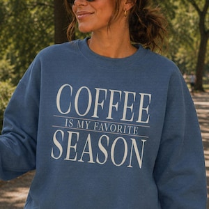 May include: A blue crewneck sweatshirt featuring the white text COFFEE IS MY FAVORITE SEASON printed on the front. The long-sleeved pullover has a ribbed neckline, cuffs, and hem, and is paired with dark brown shorts and black sunglasses.