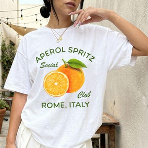 May include: White t-shirt with the text "APEROL SPRITZ Social Club ROME, ITALY" in green. The shirt features an orange graphic. The person is wearing a white pleated skirt.