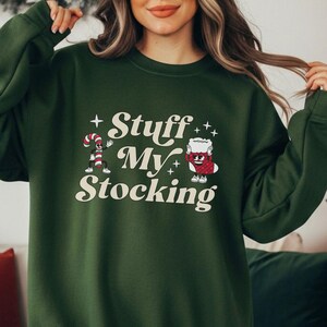 May include: A dark green sweatshirt featuring the phrase "Stuff My Stocking" in a retro font. The design includes cartoon illustrations of a candy cane and a mug of hot cocoa. The sweatshirt is a crew neck style, perfect for the festive season.