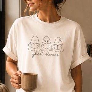 以下が含まれることがあります： オフホワイトの半袖Tシャツ。本を読む3体の幽霊の黒い線画が描かれています。1体はマグカップを持ち、1体は眼鏡をかけ、もう1体は小さなリボンをつけています。幽霊の下には筆記体で「ghost stories」という文字がプリントされています。