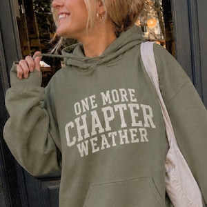 May include: An olive green hooded sweatshirt featuring a front kangaroo pocket and the text "ONE MORE CHAPTER WEATHER" printed in a cream-colored collegiate-style font across the chest. The garment includes a matching drawstring hood.