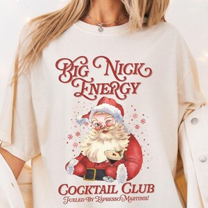 Puede incluir: Camiseta color crema con un dise&ntilde;o de Pap&aacute; Noel y el texto "Big Nick Energy Cocktail Club Fueled by Espresso Martinis!". La ilustraci&oacute;n de Pap&aacute; Noel es roja y blanca, con copos de nieve.