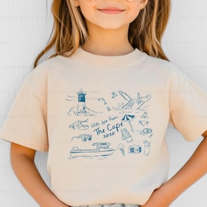 May include: A young person wearing heart-shaped sunglasses and a beige t-shirt. The t-shirt features a blue illustration of a lighthouse, whale, boat, and other beach-themed elements, with the text "With love from The Cape 2020."