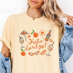 May include: A yellow t-shirt with a graphic design of a cocktail glass, orange slices, sunglasses, and the text "Feelin' Spritzy!"