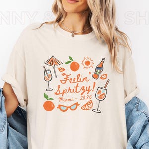 May include: A white t-shirt with a graphic design featuring a cocktail glass, orange slices, sunglasses, and the text "Feelin' Spritzy! Miami - 2025".
