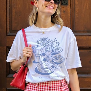May include: White t-shirt featuring a blue illustration of a coffee pot, a cup of coffee, pastries, and flowers. The text "Bon Appetit Patisserie" is printed on the shirt. The model is wearing a red and white gingham skirt and a red baseball cap.