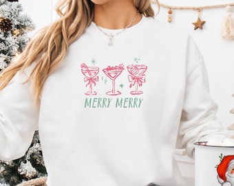 Könnte beinhalten: Weißes Sweatshirt mit einem rosa und grünen Aufdruck von drei Cocktailgläsern mit Schleifen und den Worten "MERRY MERRY". Das Sweatshirt hat lange Ärmel. Eine weiße Tasse mit einem Weihnachtsmann-Motiv wird in der Hand gehalten.