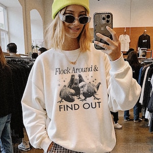 May include: White sweatshirt with a black graphic of a flock of birds and the text "Flock Around & Find Out". The model is wearing a bright green beanie and sunglasses.