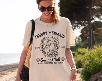 May include: A woman wearing a light beige t-shirt with a black graphic of a manatee in a floral wreath. The text on the shirt reads "Chubby Mermaid Social Club Est. 1998 Where Chill is the New Thrill".