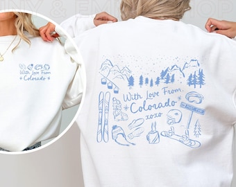 May include: White sweatshirt with a blue graphic design on the back. The graphic features a mountain scene with ski equipment, trees, and the text "With Love From *Colorado* xoxo". The front of the sweatshirt has the text "With Love From *Colorado*" in blue.