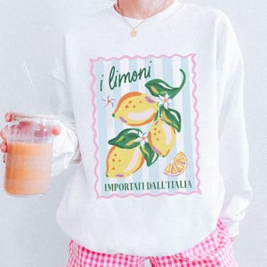 May include: White sweatshirt featuring a lemon graphic with the text "i limoni IMPORTATI DALL'ITALIA". The graphic includes yellow lemons, green leaves, and white flowers. The sweatshirt is paired with pink and white checkered shorts.