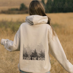 May include: A beige hoodie with a forest silhouette and the text "Leave the Road" printed on the back. The hoodie is worn by a person standing in a field.