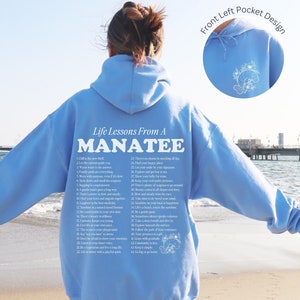K&ouml;nnte beinhalten: Ein hellblauer Hoodie mit einem wei&szlig;en Grafikdruck eines Manati und einer Liste von Lebenslektionen eines Manati. Der Text auf dem Hoodie lautet "Life Lessons From A MANATEE" und enth&auml;lt 22 Lebenslektionen. Der Hoodie hat eine Fronttasche mit einem kleinen gestickten Manati-Design.