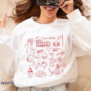 May include: White sweatshirt with a red graphic design of Italian icons, including the Colosseum, a gondola, a Vespa scooter, and the text "Ciao Italia".
