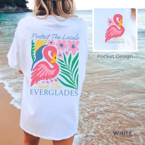May include: White t-shirt with a pink flamingo and floral design. The text "Protect The Locals Save The EVERGLADES" is printed on the back of the shirt. A smaller version of the flamingo design is printed on the front pocket of the shirt.