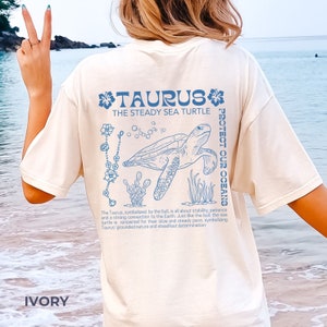 May include: Ivory t-shirt featuring a blue sea turtle graphic and the text "TAURUS THE STEADY SEA TURTLE PROTECT OUR OCEANS". The text below the graphic reads "The Taurus, symbolised by the bull, is all about stability, patience and a strong connection to the Earth. Just like the bull, the sea turtle is renowned for their slow and steady pace, symbolising Taurus' grounded nature and steadfast determination."