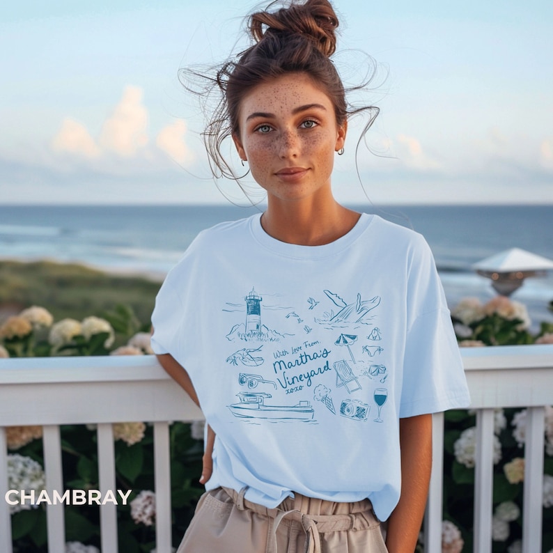 May include: A light blue t-shirt with a graphic design featuring a lighthouse, a whale, a boat, and the text "With love from Martha's Vineyard 2023".