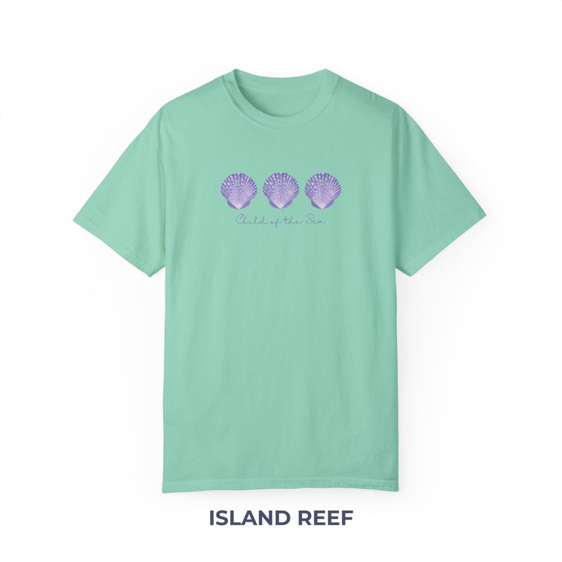 Pu&ograve; includere: Una maglietta verde chiaro con una grafica di tre conchiglie viola e il testo "Child of the Sea". La maglietta &egrave; di Island Reef.