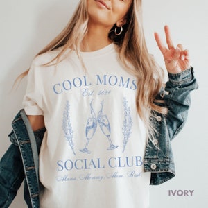 May include: White t-shirt with blue text that reads "Cool Moms Est. 2024 Social Club Mama, Mommy, Mom, Bruh."  The shirt features a graphic of two champagne glasses clinking together.