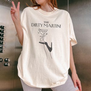 May include: A cream colored t-shirt with the text "The Dirty Martini" printed in black. The graphic features a martini glass with olives and a black high heel shoe.