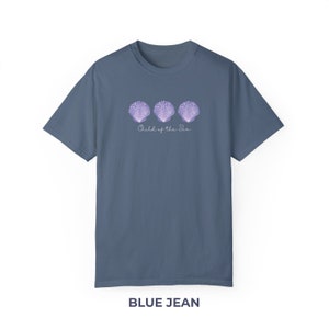 Pu&ograve; includere: Una t-shirt blu jeans con una grafica di tre conchiglie viola e il testo "Child of the Sea".
