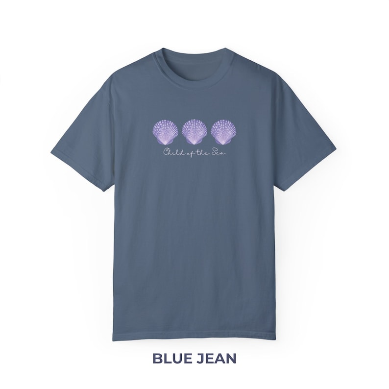 Pu&ograve; includere: Una t-shirt blu jeans con una grafica di tre conchiglie viola e il testo "Child of the Sea".