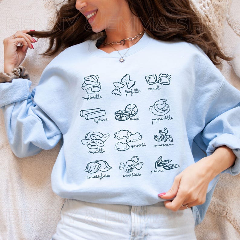 May include: Light blue sweatshirt with a graphic of different types of pasta, including tagliatelle, farfalle, ravioli, rigatoni, cavatelli, pappardelle, gnocchi, macaroni, conchigliette, orecchiette, and penne.