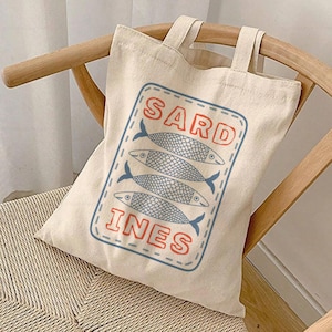 May include: A white canvas tote bag with a blue and red graphic of four sardines and the text "SARDINES".