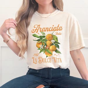 May include: Cream-colored t-shirt featuring an orange graphic of oranges with green leaves and the text "Aranciata Italia 1987 La Dolce Vita" in orange script. The shirt has a relaxed fit.