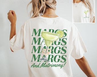 Może przedstawiać: Kremowa koszulka z zieloną grafiką margarity i napisem "MARGS MARGS MARGS And Matrimony!" z tyłu. Mniejsza wersja grafiki margarity znajduje się z przodu. Koszulka ma luźny krój.