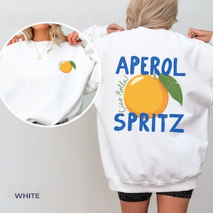 May include: White sweatshirt with a blue and orange graphic on the back that says "Aperol Spritz" with the text "Ciao Bella!" written vertically next to the orange. The front of the sweatshirt has a small orange graphic with the text "Ciao Bella!" written vertically next to the orange.