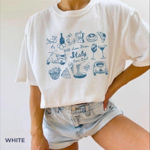 May include: White t-shirt featuring a blue graphic print of Italian icons, including a scooter, pizza, pasta, wine, and the text "With Love From Italy Ciao, Ciao!"
