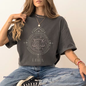 Könnte beinhalten: Ein dunkelgraues T-Shirt mit einem Grafikdruck eines Libra-Sternzeichen-Symbols in einem Kreis mit dem Wort "LIBRA" darunter. Das Symbol ist eine stilisierte Waage mit einem Stern darüber.