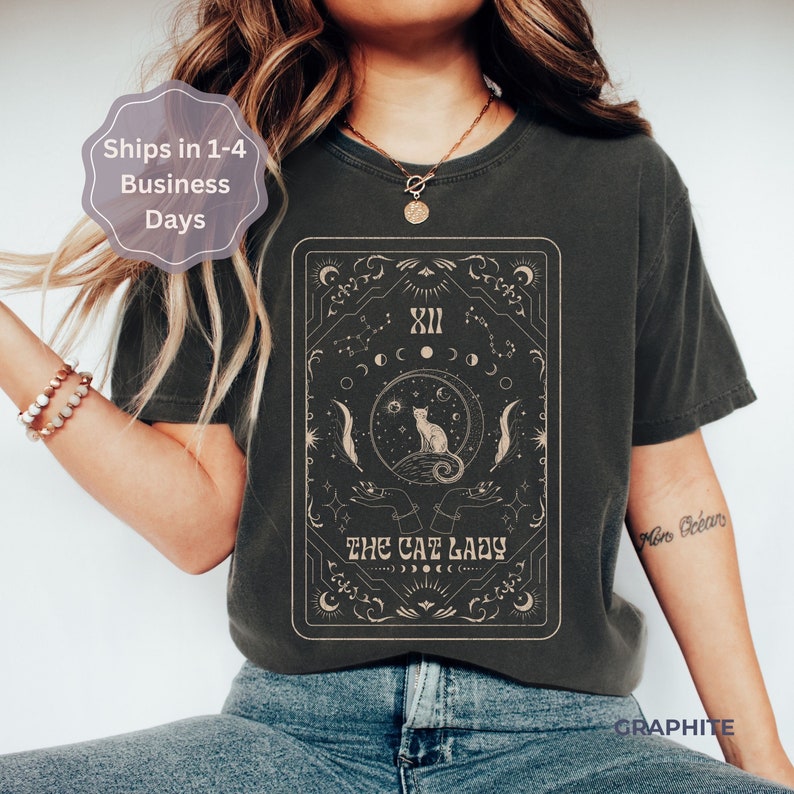 Pode incluir: Uma camiseta cinza escura com uma estampa gr&aacute;fica de uma carta de tar&ocirc; com um gato sentado em uma lua crescente. A carta &eacute; cercada por estrelas e luas. O texto "The Cat Lady" est&aacute; impresso na carta.