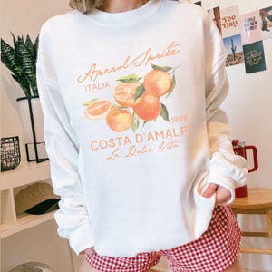 May include: White sweatshirt with the text "Aperol Spritz Italia" and a watercolor illustration of oranges. The design includes the words "Costa D'Amalfi" and "La Dolce Vita" in a script font. The sweatshirt has long sleeves.