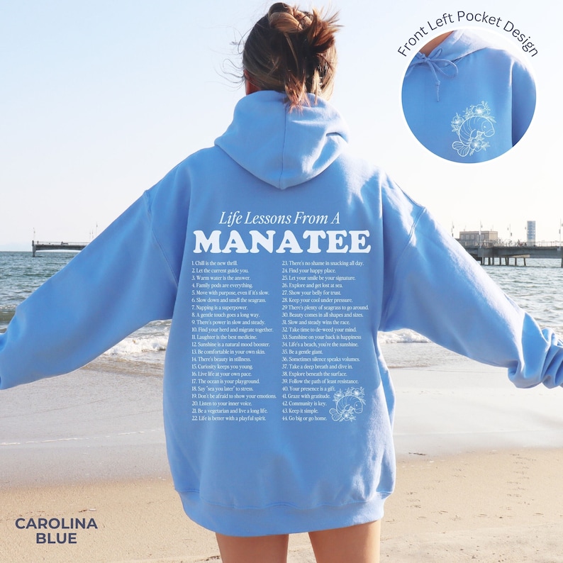 K&ouml;nnte beinhalten: Ein hellblauer Hoodie mit einem wei&szlig;en Grafikdruck eines Manati und einer Liste von Lebenslektionen eines Manati. Der Text auf dem Hoodie lautet "Life Lessons From A MANATEE" und enth&auml;lt 22 Lebenslektionen. Der Hoodie hat eine Fronttasche mit einem kleinen gestickten Manati-Design.