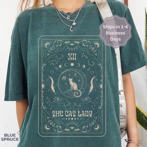 Pode incluir: Camiseta azul turquesa com uma ilustra&ccedil;&atilde;o vintage de um gato sentado em uma lua crescente com estrelas e s&iacute;mbolos celestiais. O texto "The Cat Lady" est&aacute; impresso abaixo do gato.