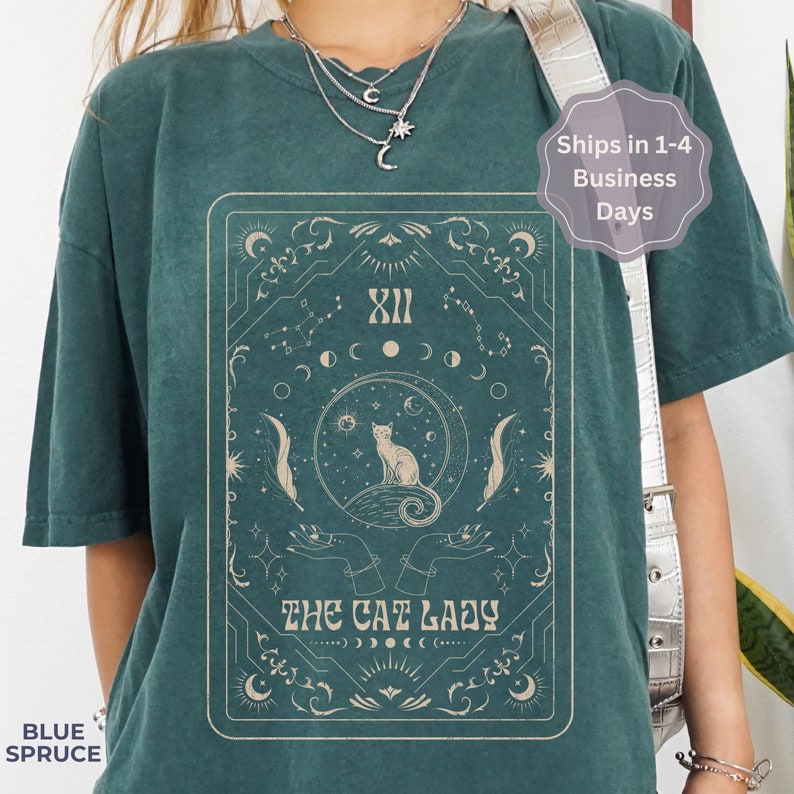Pode incluir: Camiseta azul turquesa com uma ilustra&ccedil;&atilde;o vintage de um gato sentado em uma lua crescente com estrelas e s&iacute;mbolos celestiais. O texto "The Cat Lady" est&aacute; impresso abaixo do gato.