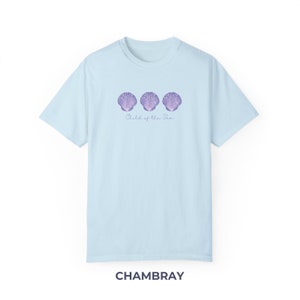 Pu&ograve; includere: Una t-shirt blu chiaro con tre conchiglie viola e il testo "Child of the Sea" stampato sul davanti. La t-shirt &egrave; realizzata in tessuto chambray morbido e confortevole.