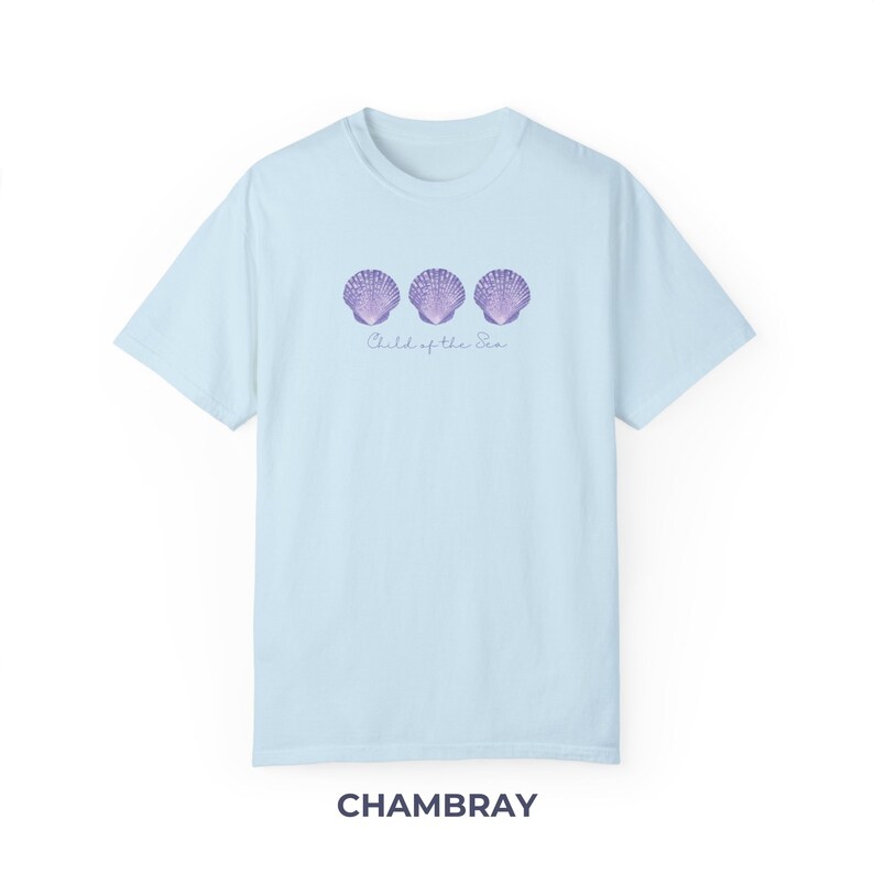 Pu&ograve; includere: Una t-shirt blu chiaro con tre conchiglie viola e il testo "Child of the Sea" stampato sul davanti. La t-shirt &egrave; realizzata in tessuto chambray morbido e confortevole.