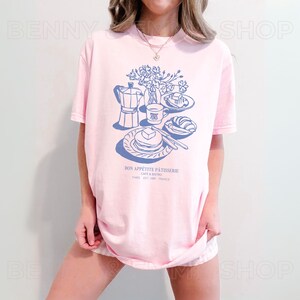 May include: A pink t-shirt featuring a blue illustration of a breakfast scene. The illustration includes a coffee pot, a cup of coffee, a plate of pancakes, a croissant, and a bouquet of flowers. The text "Bon App&eacute;tit P&acirc;tisserie, Caf&eacute; & Bistro, Paris Est. 1988 France" is printed below the illustration.