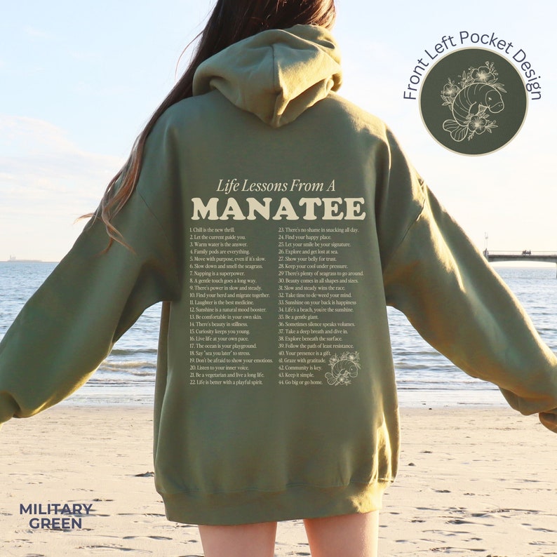 K&ouml;nnte beinhalten: Ein gr&uuml;ner Kapuzenpullover mit einem grafischen Aufdruck eines Seekuhs und dem Text "Life Lessons From A Manatee" mit einer Liste von 44 Lebenslektionen. Der Text ist in einem hellen Cremeton gehalten. Der Pullover hat eine Tasche auf der linken Seite mit einer kleinen Grafik eines Seekuhs.